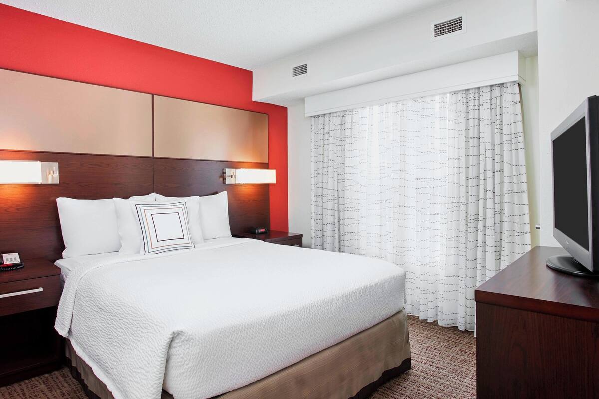 A hotel room with a large bed, a wooden headboard, a bedside table with a lamp, a television, and a window with curtains.