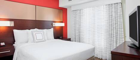 Une chambre d’hôtel avec un grand lit, une tête de lit en bois, une table de chevet avec une lampe, une télévision et une fenêtre avec des rideaux.