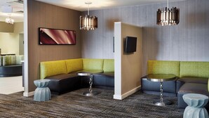 Eine moderne Hotellobby mit einer Eckcouch, einem kleinen runden Tisch und einem an der Wand montierten Flachbildfernseher.
