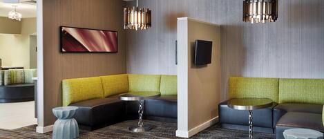 A modern hotel lobby with a sectional sofa, a small round table, and a flat-screen TV mounted on the wall.