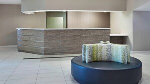 Ein Empfangsbereich mit modernem Design, ausgestattet mit einer Rezeption, einer Sitzecke mit einem gemusterten Pouf und einer dekorativen Leuchte.