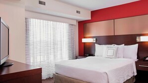 Ein Hotelzimmer mit einem großen Bett, einem Fernseher auf einer Holzkommode, einem Nachttisch mit Lampe und einem Fenster mit durchsichtigen Vorhängen.