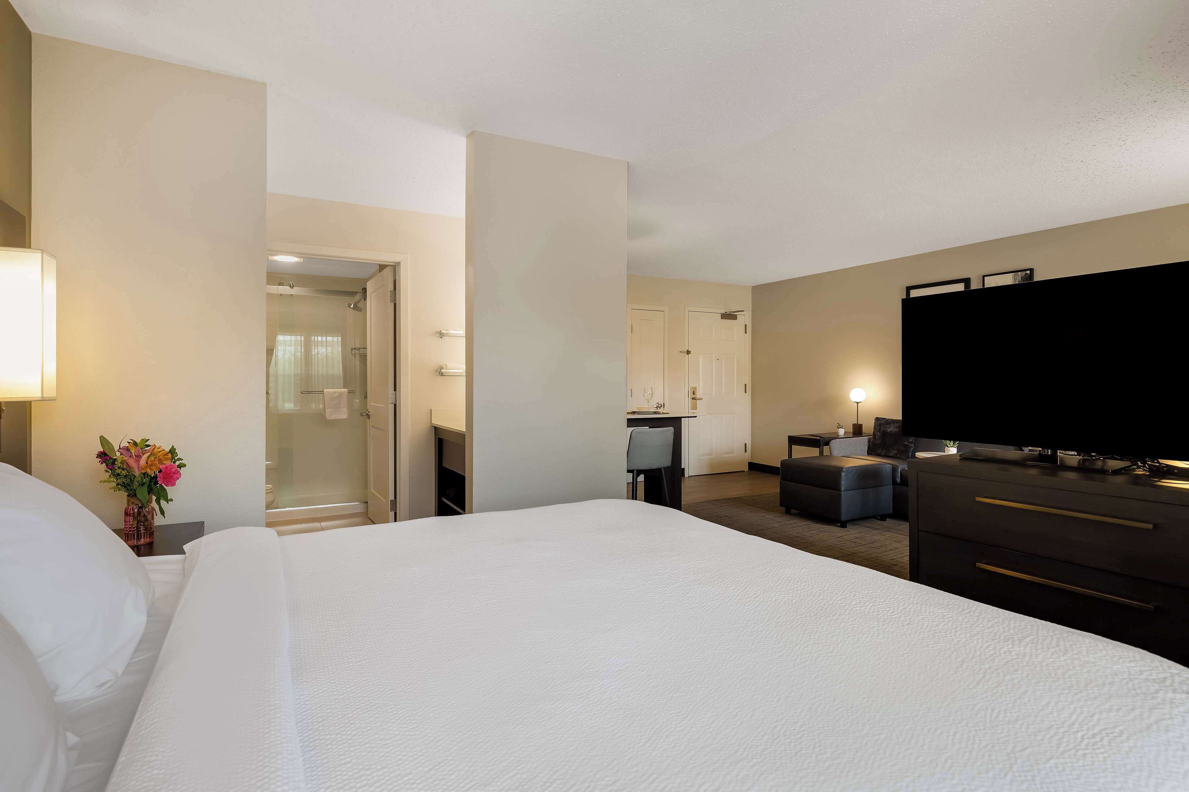 Habitación de hotel con cama, escritorio, televisor y baño visible a través de una puerta abierta.