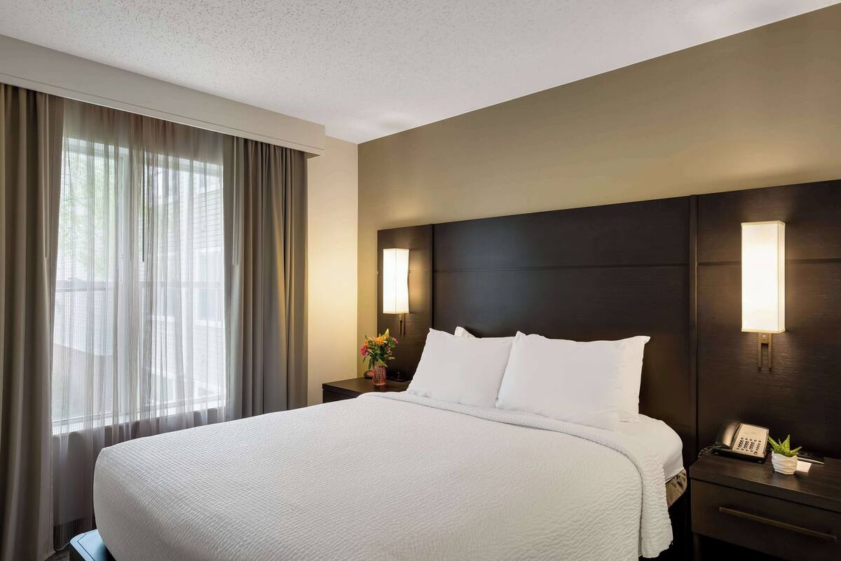 Ein Hotelzimmer mit einem großen Bett, einem Nachttisch mit Lampe und einem Fenster mit Vorhängen.