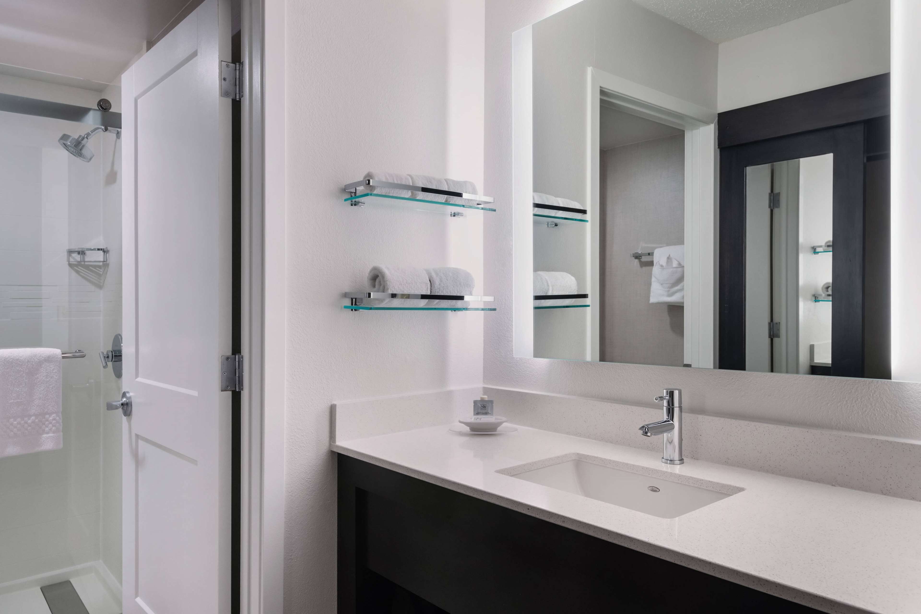 Une salle de bain moderne avec un lavabo blanc, un grand miroir et des étagères en verre où sont rangées les serviettes.