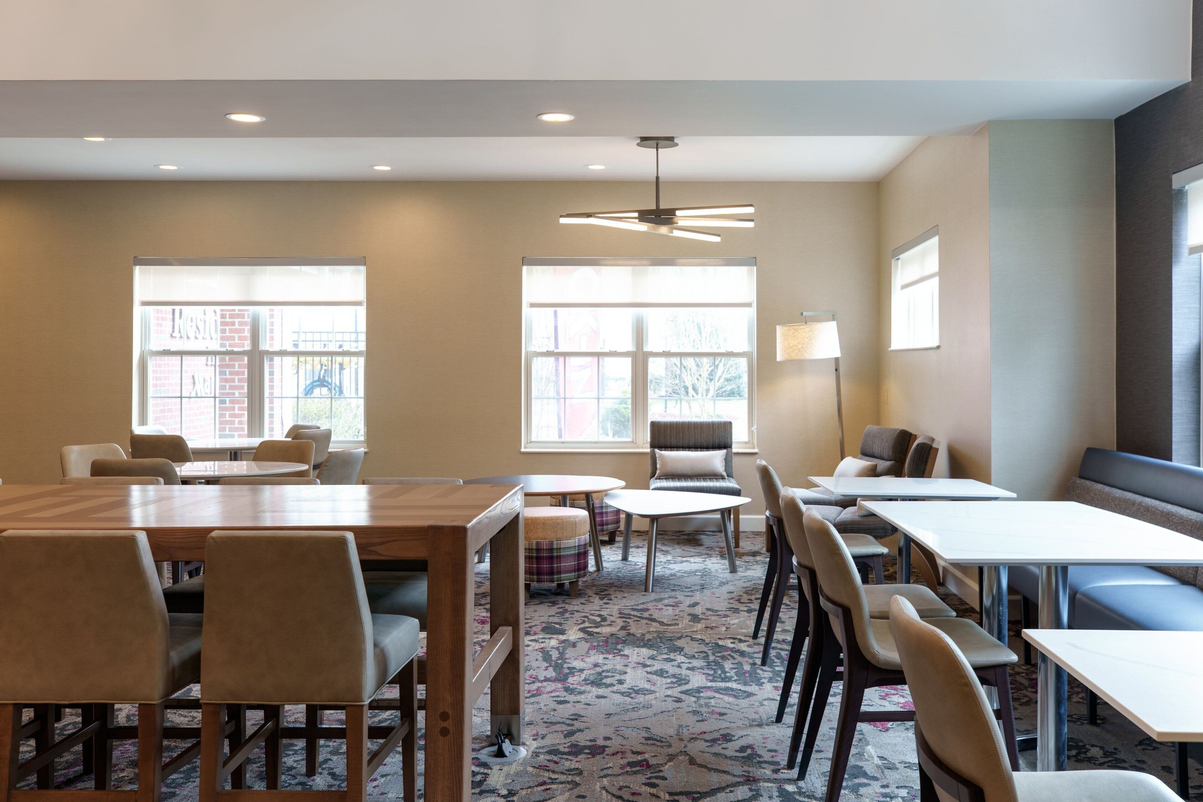 A modern conference room with a large wooden table, several chairs, a small round table, and a sofa. There are large windows providing natural light and a view of the outside.