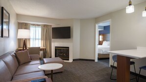 A hotel room with a sofa, a small round table, a flat-screen TV mounted on the wall, a fireplace, and a separate bedroom with a bed and a nightstand. - Residence Inn By Marriott Columbia (Ellicott City)