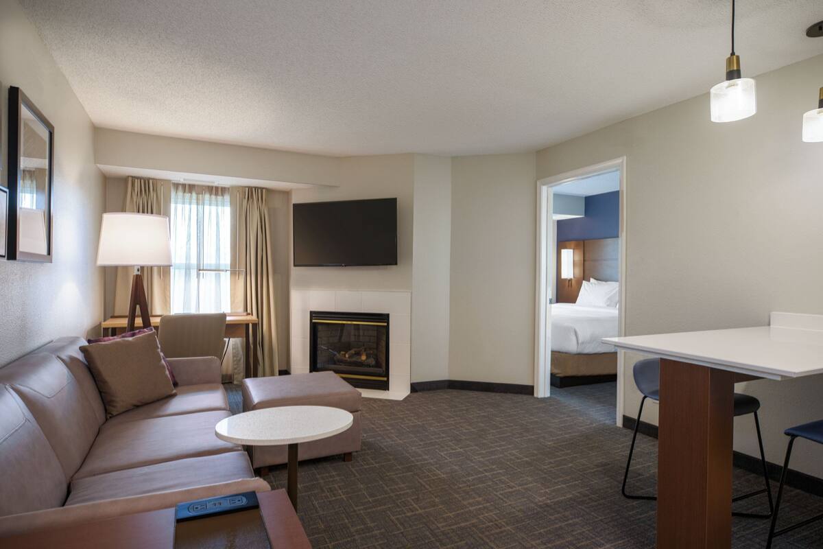 A hotel room with a sofa, a small round table, a flat-screen TV mounted on the wall, a fireplace, and a separate bedroom with a bed and a nightstand.