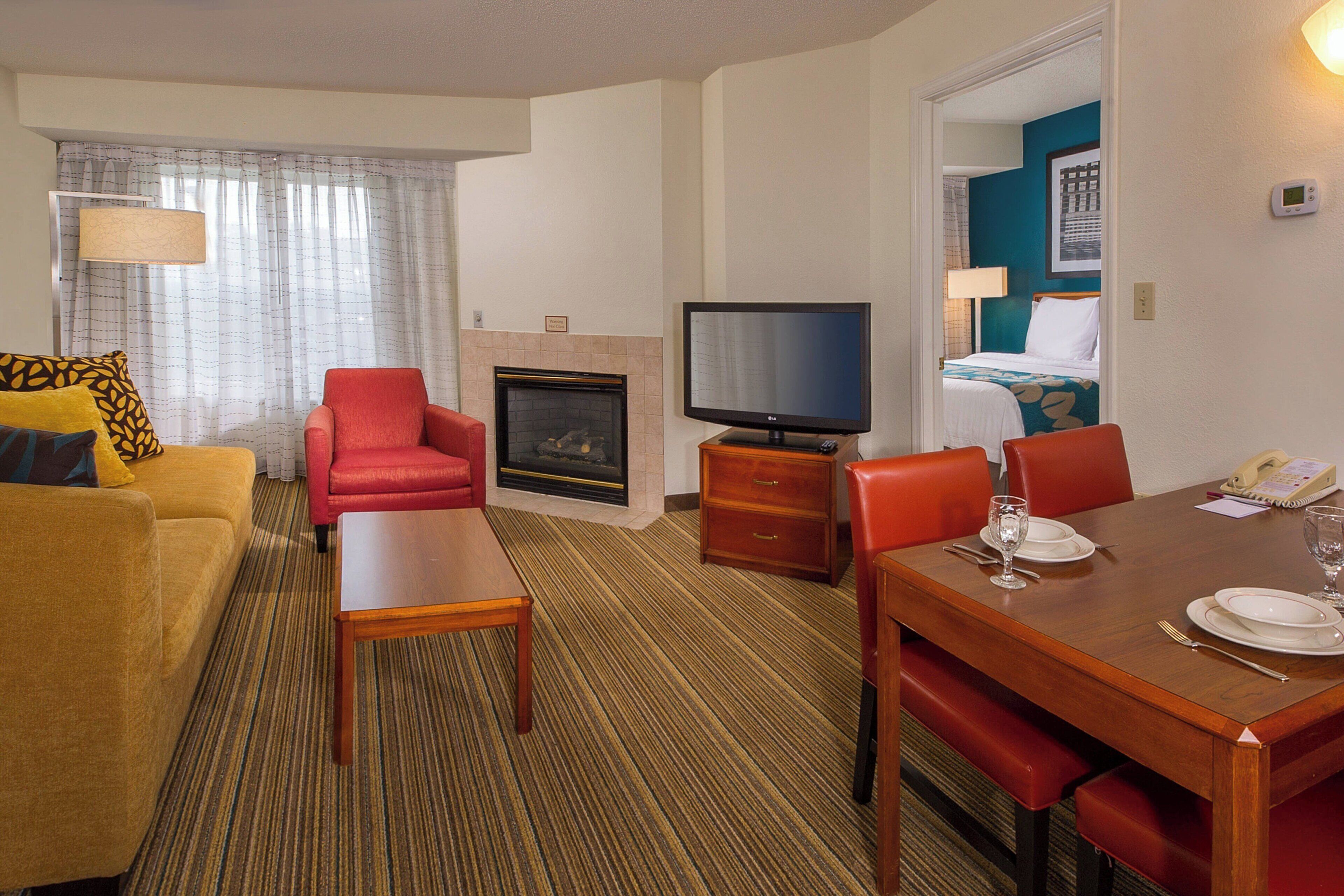 A hotel room with a sofa, a red armchair, a wooden table, a television, and a fireplace.