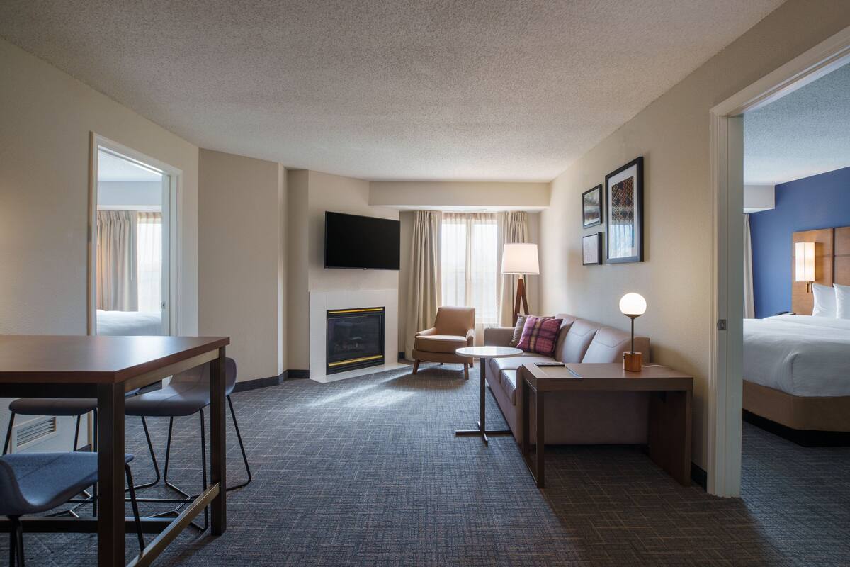 A hotel room with a flat-screen TV, a fireplace, a sofa, a dining table with chairs, and a bed.