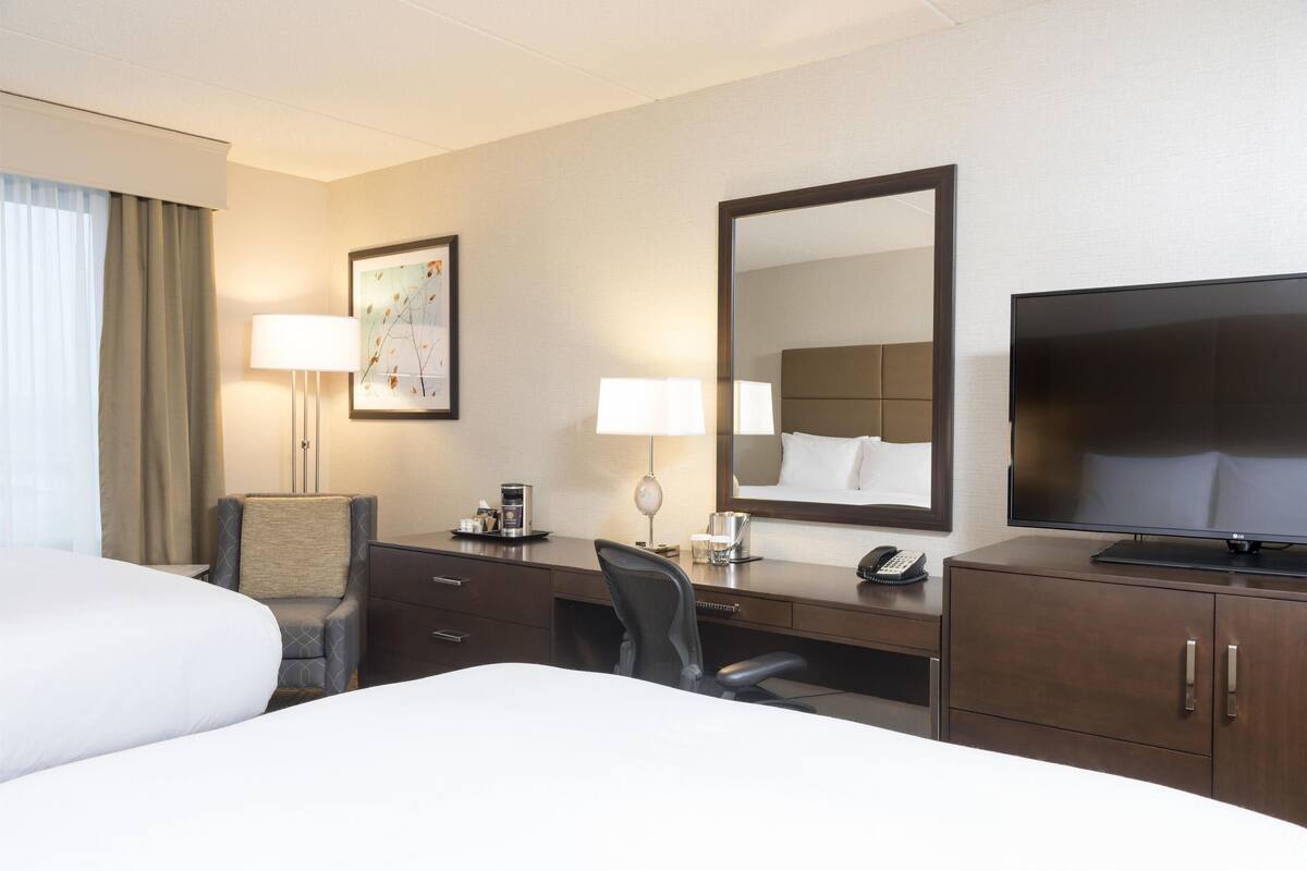 A hotel room with a bed, desk, chair, desk lamps, a large mirror, a television, and a telephone.