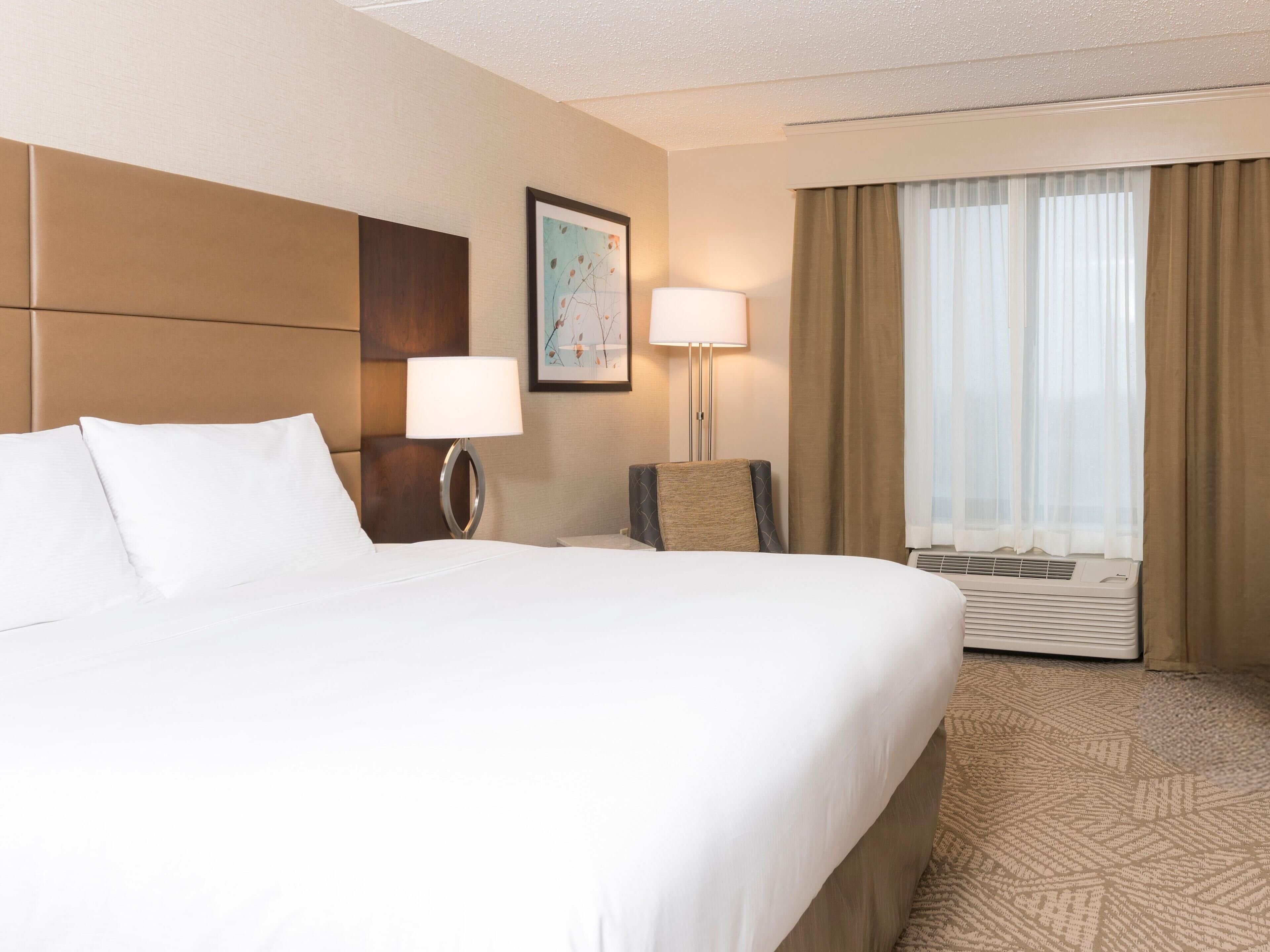 Habitación de hotel con una cama grande, un escritorio, una silla, una lámpara, un cuadro en la pared y una ventana con cortinas.