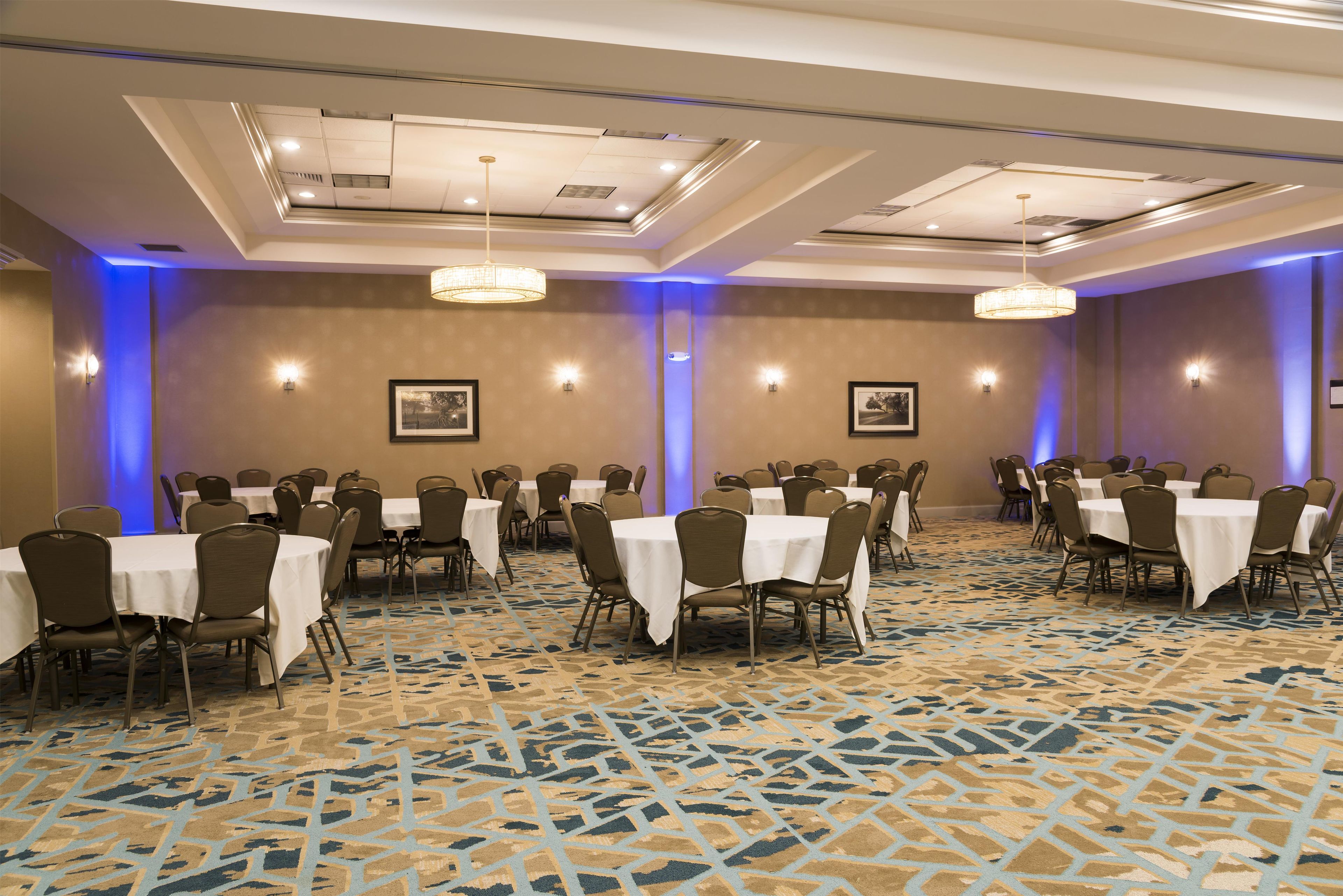 Un salón de conferencias con varias mesas y sillas, alfombra estampada e iluminación en el techo.