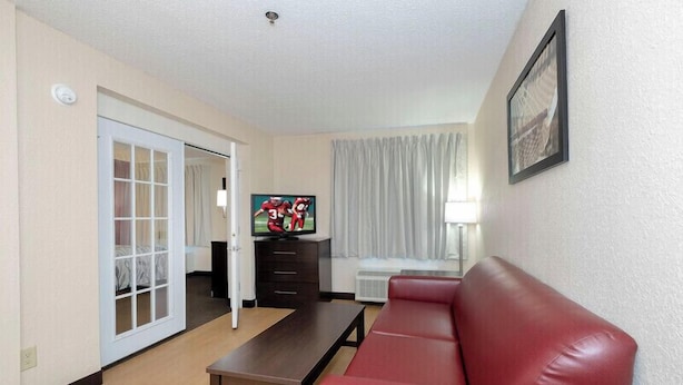 A living room with a red leather sofa, a wooden coffee table, a TV on a stand, a sliding glass door leading to a balcony, and a framed picture on the wall.