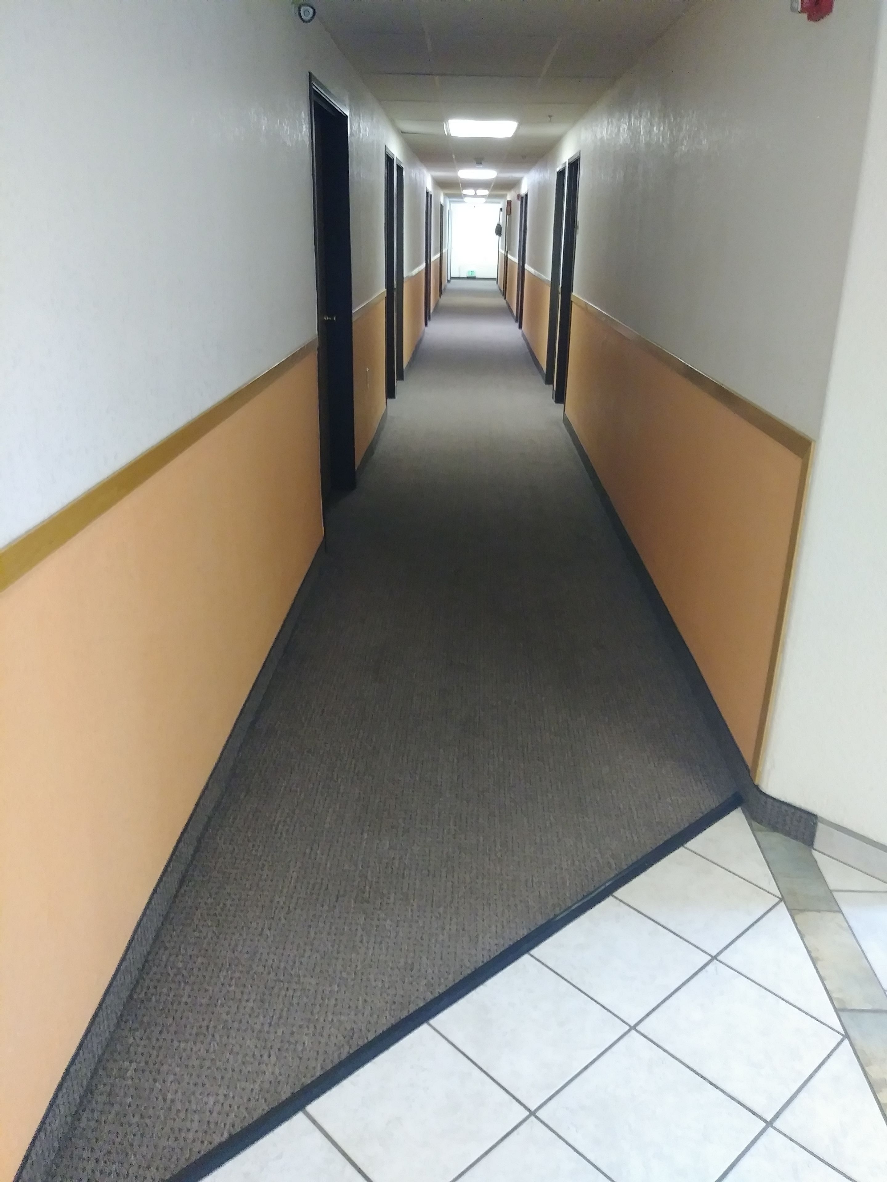 Un pasillo largo y estrecho con piso alfombrado y paredes de baldosas, que conduce a una salida luminosa al final.