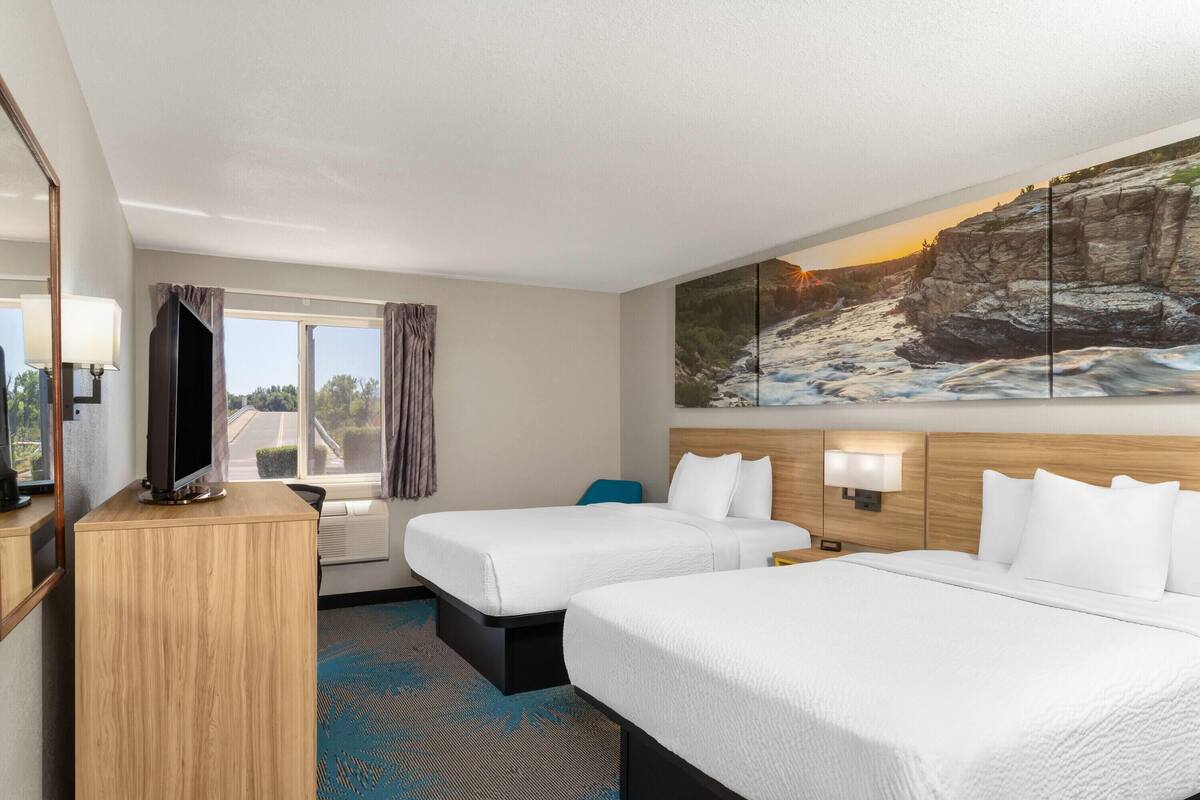 A hotel room with two beds, a television on a wooden cabinet, a large window with curtains, and a painting of a rocky landscape.
