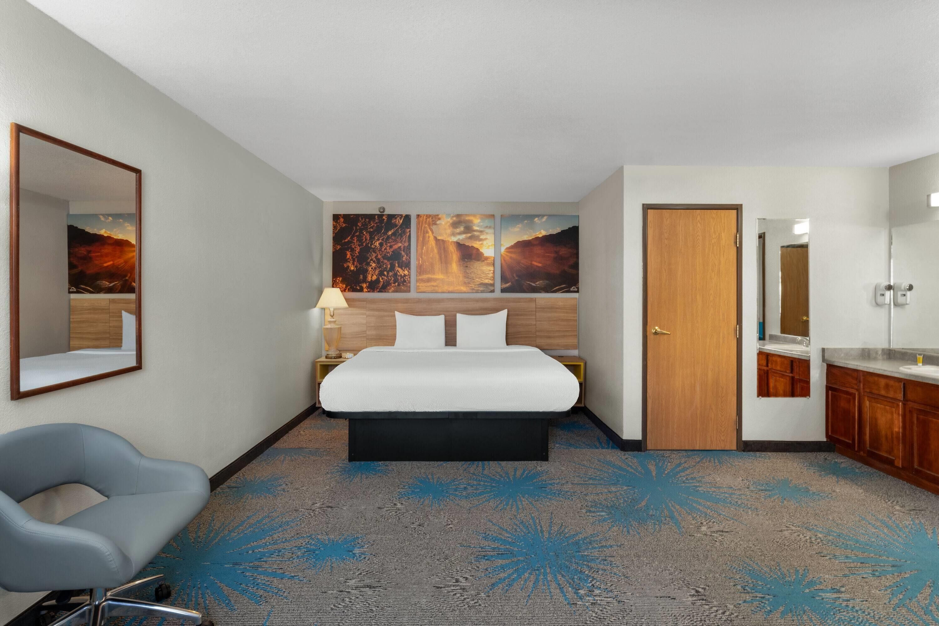 A hotel room with a bed, a chair, a desk, a mirror, and artwork on the wall.