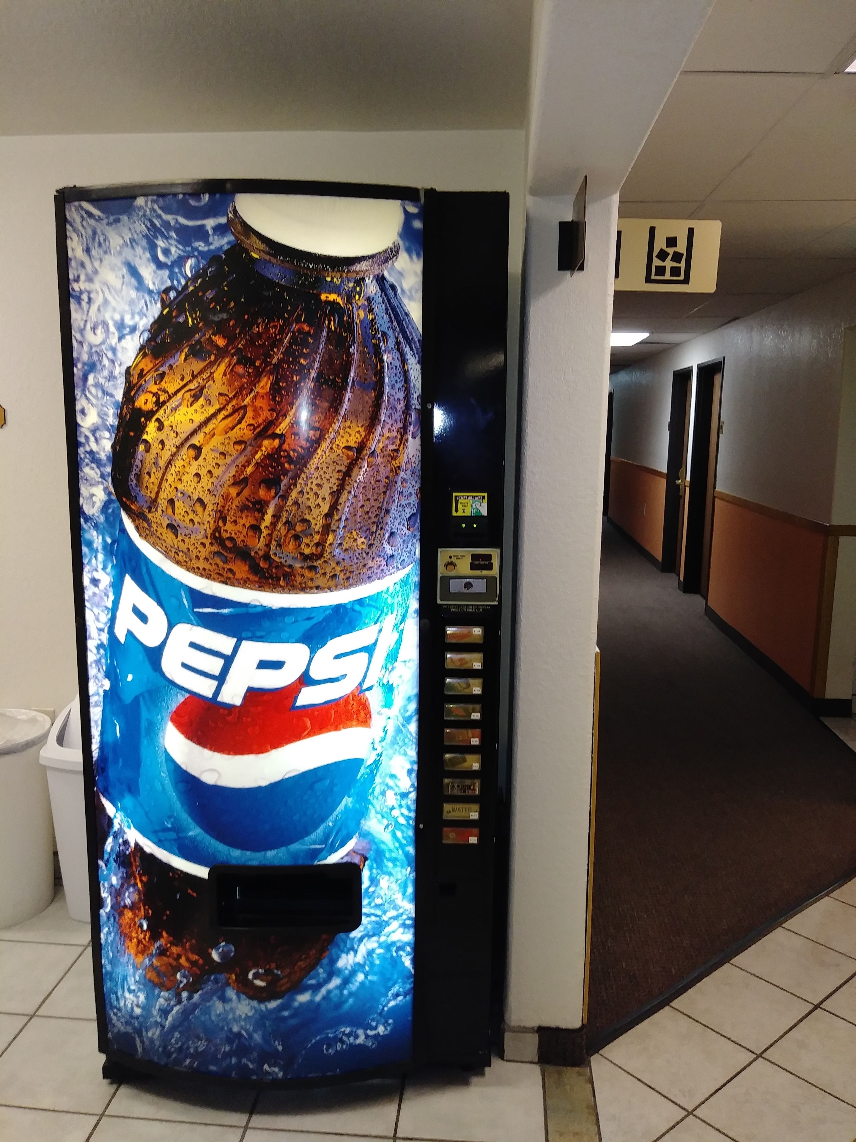 Máquina expendedora con logotipo de Pepsi, ubicada en un pasillo con suelo de baldosas y puertas.