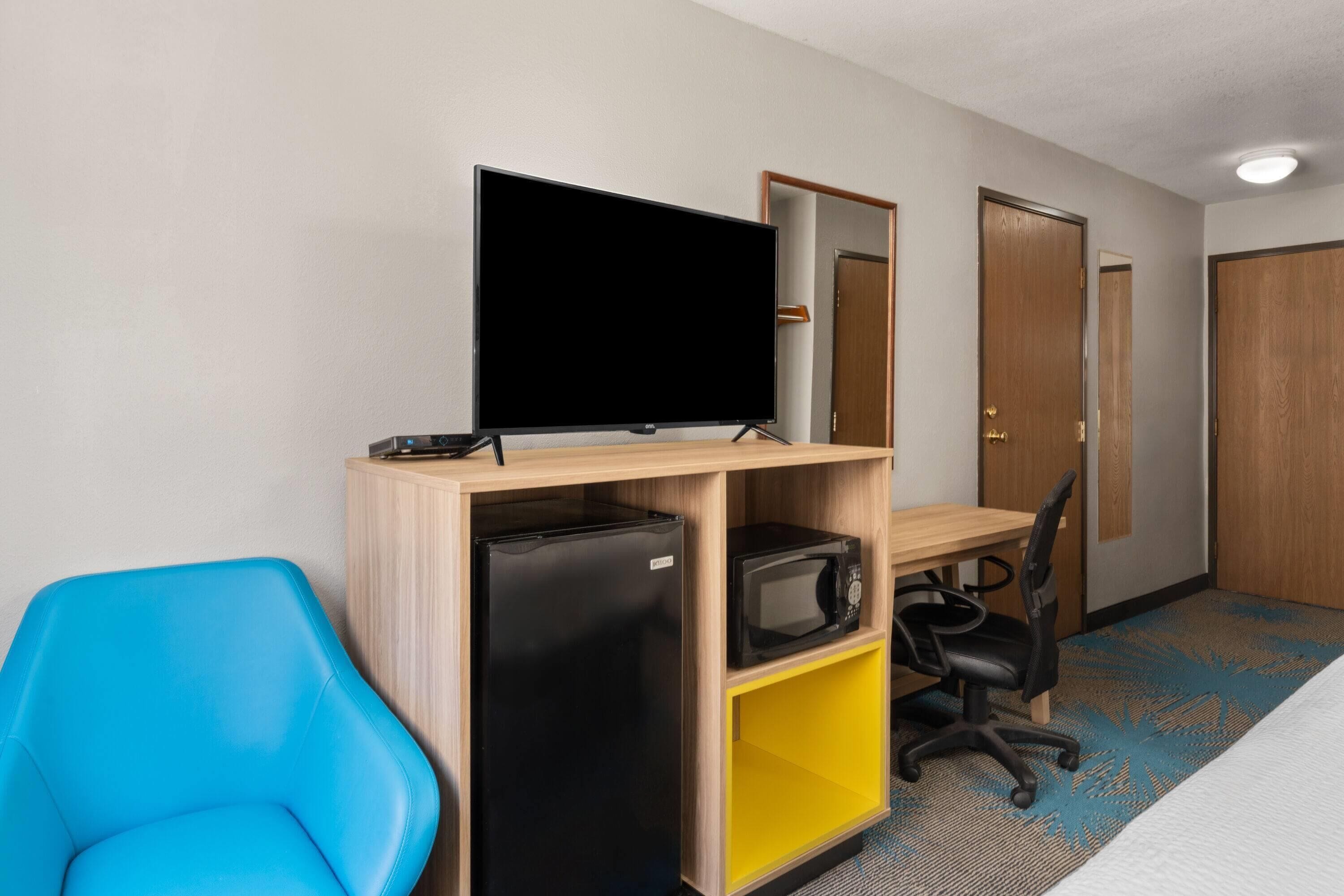 A hotel room with a bed, a desk with a flat-screen TV, a refrigerator, a microwave, a blue chair, and a door.