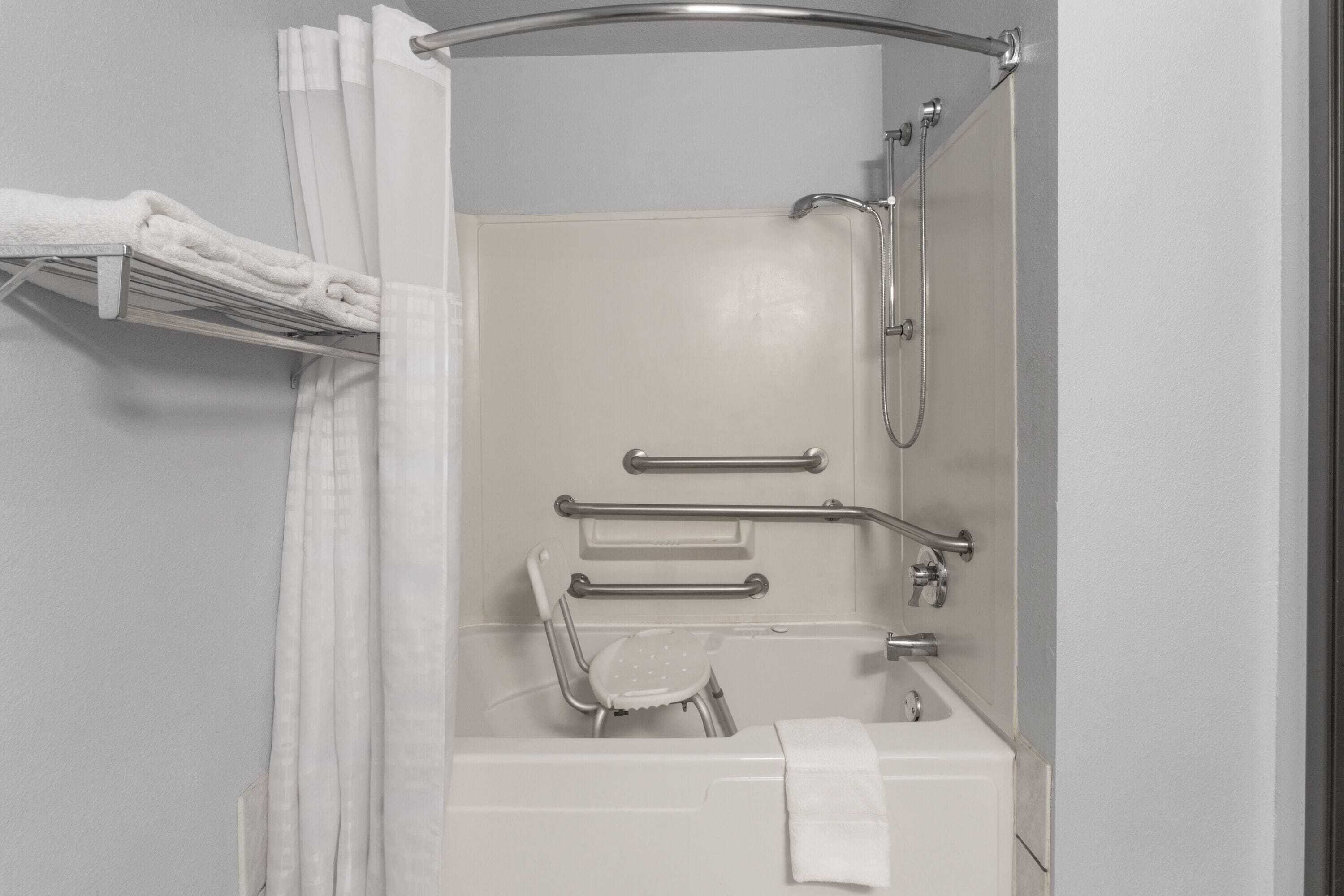 A bathroom with a shower, grab bars, a chair, and a towel rack.