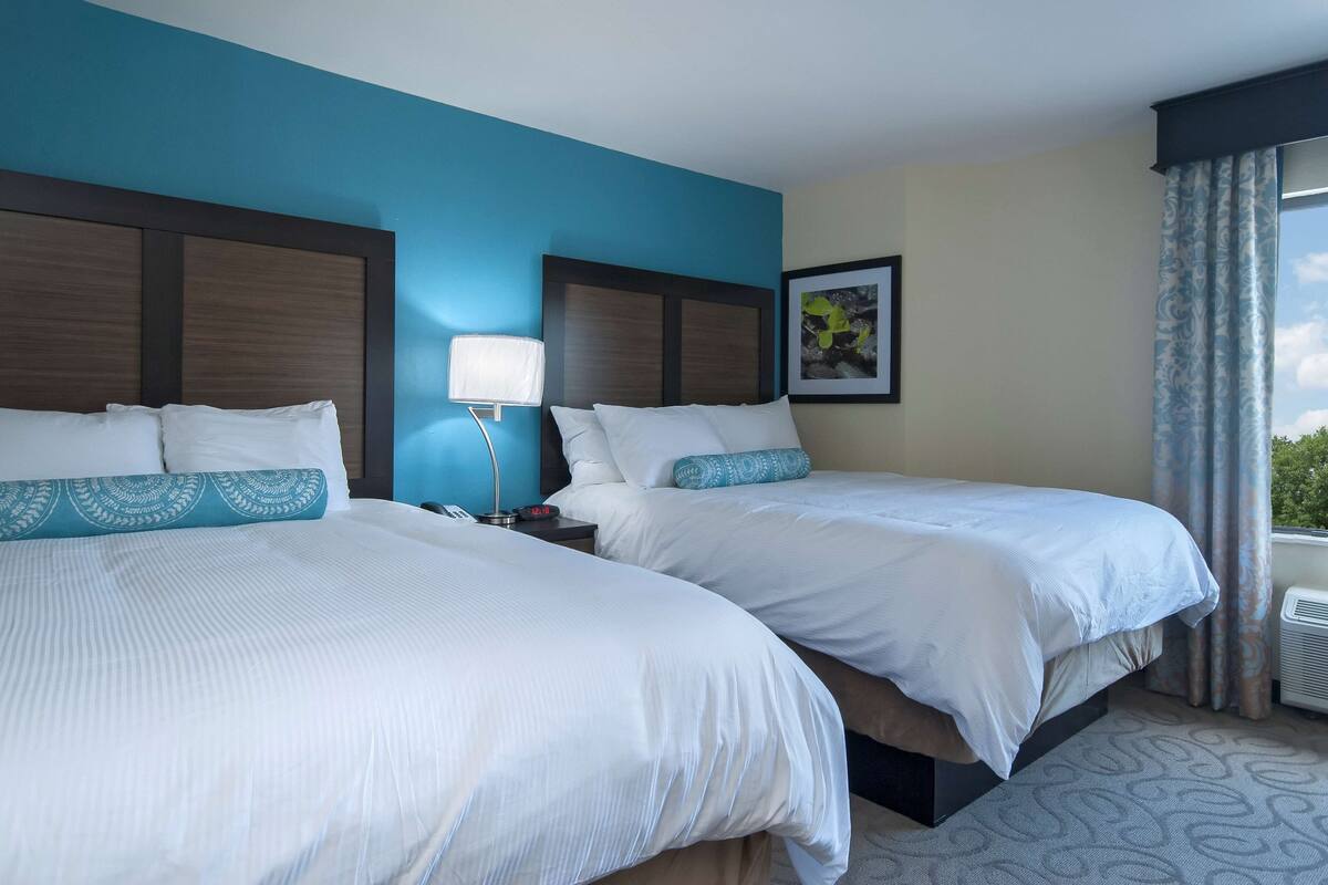 A hotel room with two beds, a nightstand with a lamp, a framed picture on the wall, and a window with curtains.
