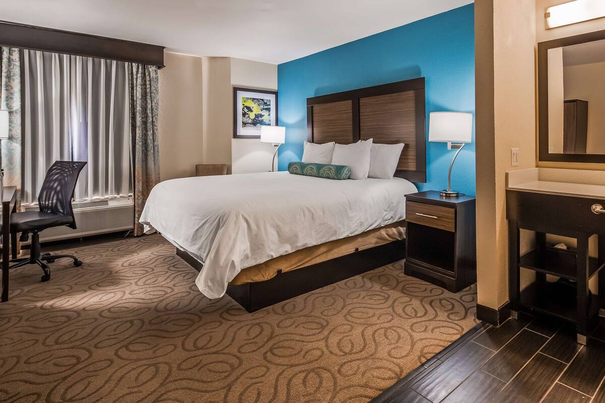 A hotel room with a large bed, a desk with a chair, a desk lamp, a nightstand with a lamp, a bathroom with a sink and mirror, and a window with curtains.