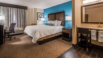 A hotel room with a large bed, a desk with a chair, a desk lamp, a nightstand with a lamp, a bathroom with a sink and mirror, and a window with curtains.