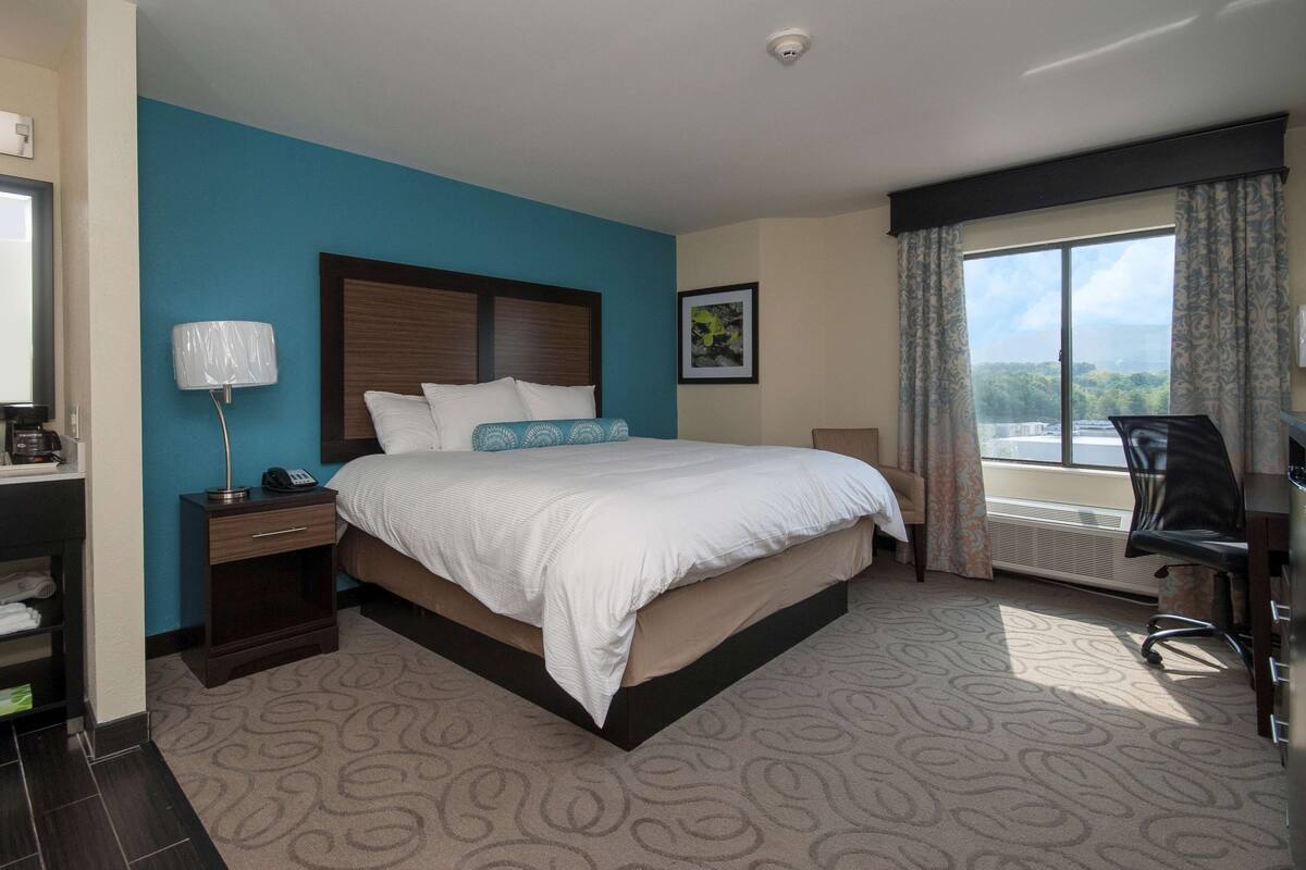 A hotel room with a large bed, a desk with a chair, a window with curtains, a framed picture on the wall, and a bathroom area with towels and toiletries.