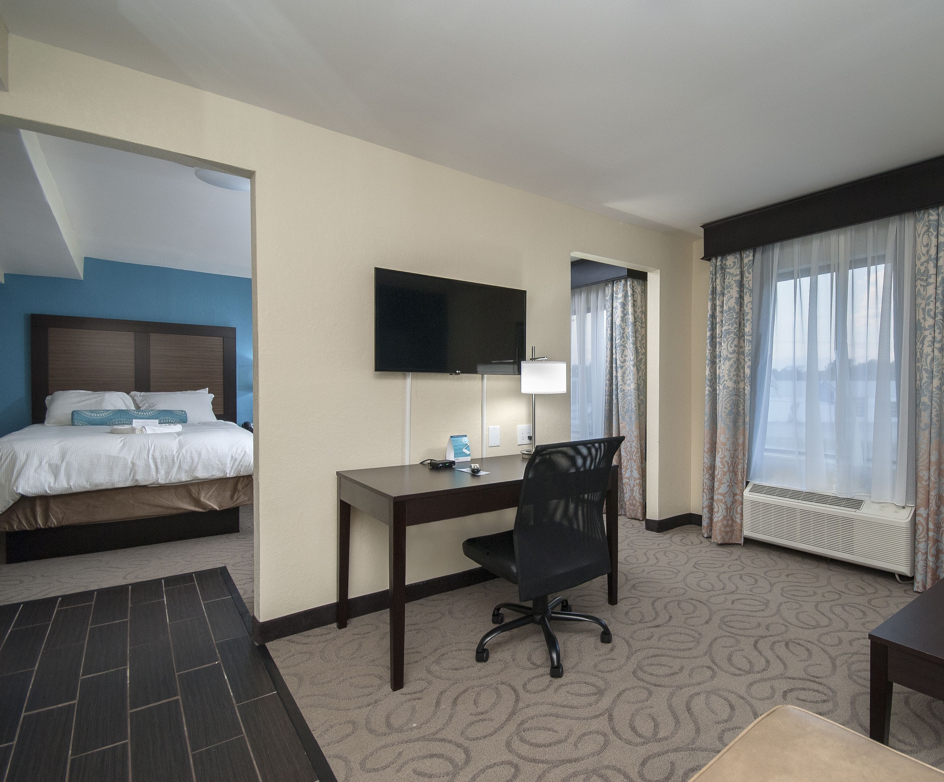 A hotel room with a bed, a desk, a chair, a TV, a window with curtains, and a radiator.