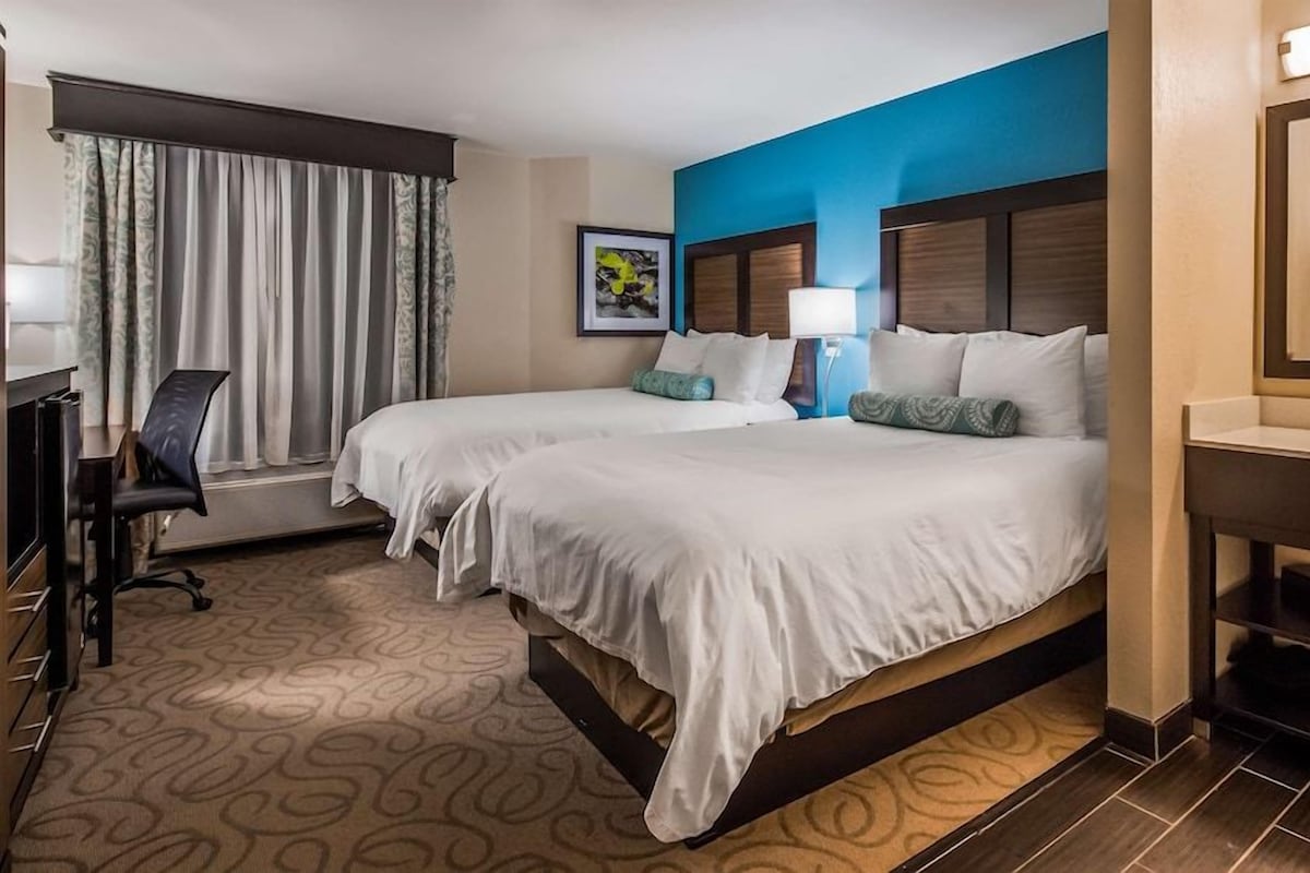 A hotel room with two beds, a desk, a chair, a bathroom with a mirror, and a window with curtains.