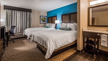 Quarto de hotel com duas camas, uma escrivaninha, uma cadeira, uma luminária de mesa, banheiro com espelho e uma janela com cortinas.
