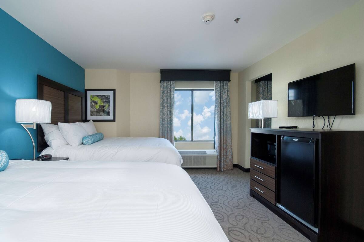 A hotel room with two beds, a TV, a desk, and a window with curtains.