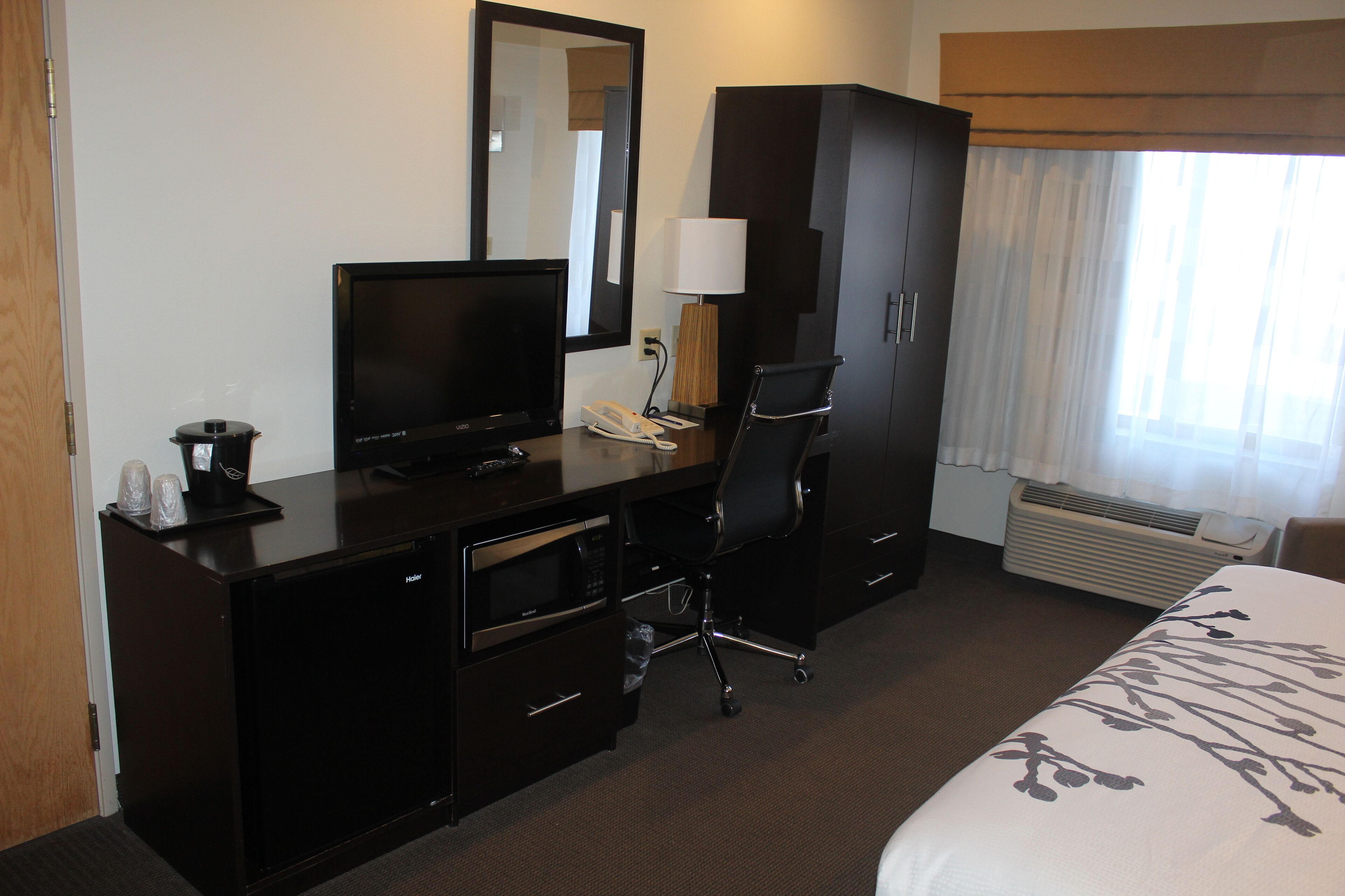 A hotel room with a bed, a desk with a computer, a chair, a television, a mirror, a wardrobe, and a window with curtains.