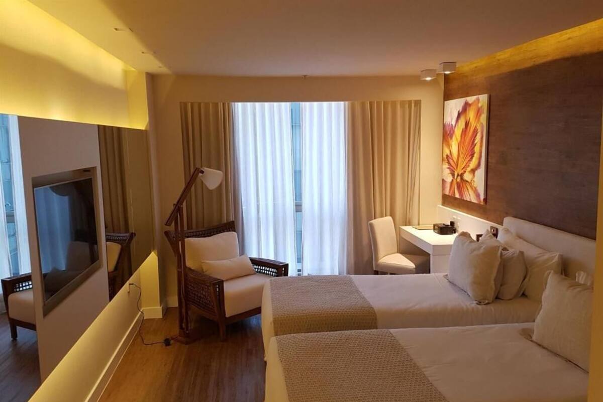 A hotel room with two beds, a desk, a chair, a TV, and a large window with curtains.