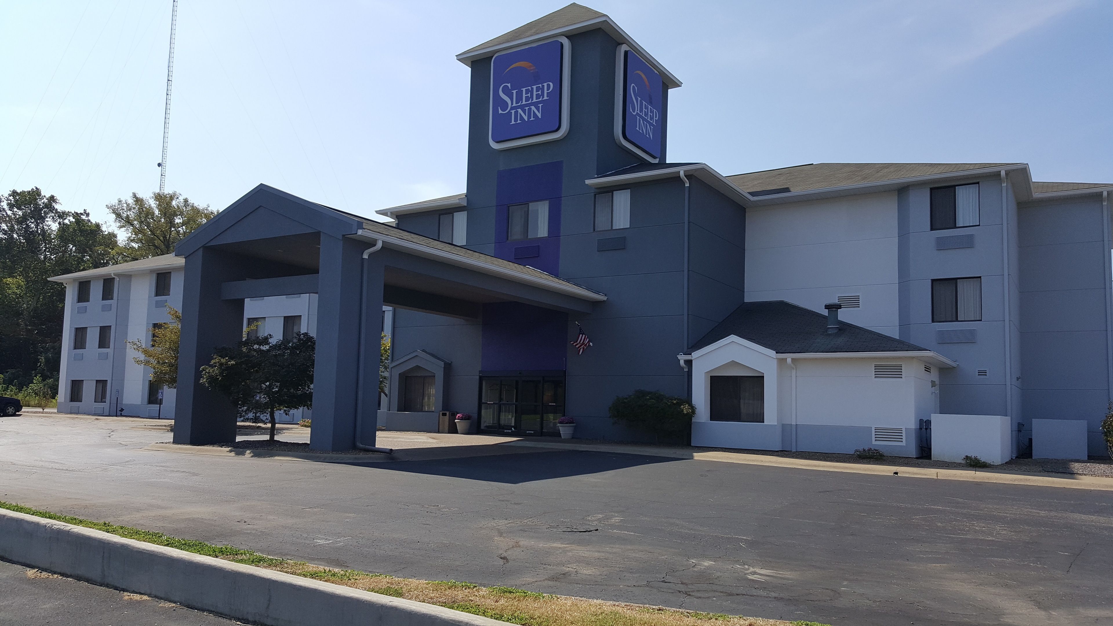 Een Sleep Inn hotel met een modern design, herkenbaar aan een opvallende toren met signage, een overdekte ingang en een parkeerplaats.