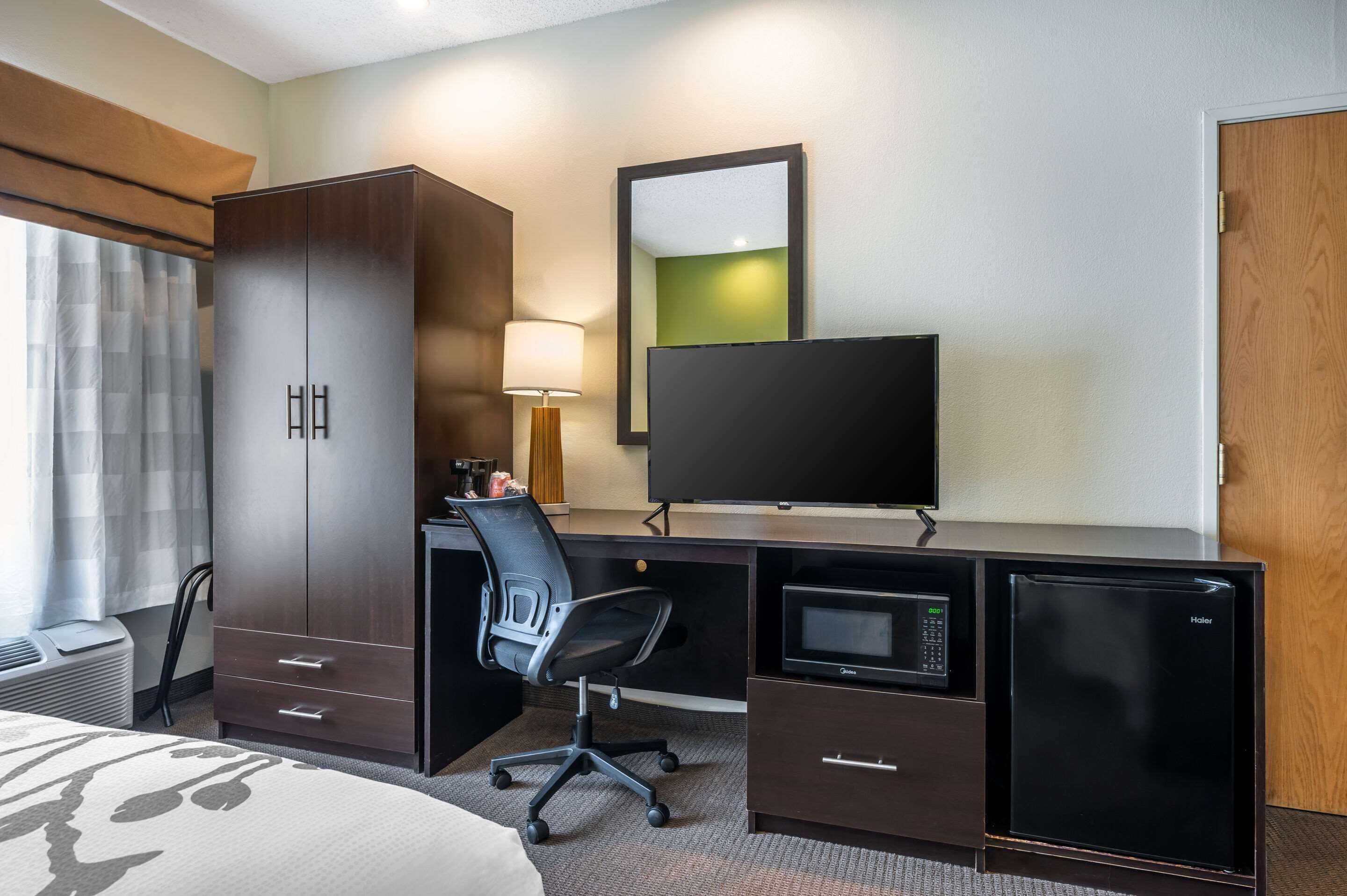 Une chambre d’hôtel comprenant un lit, un bureau avec un ordinateur et un écran, un four à micro-ondes, un réfrigérateur, une chaise, une armoire et un grand miroir.