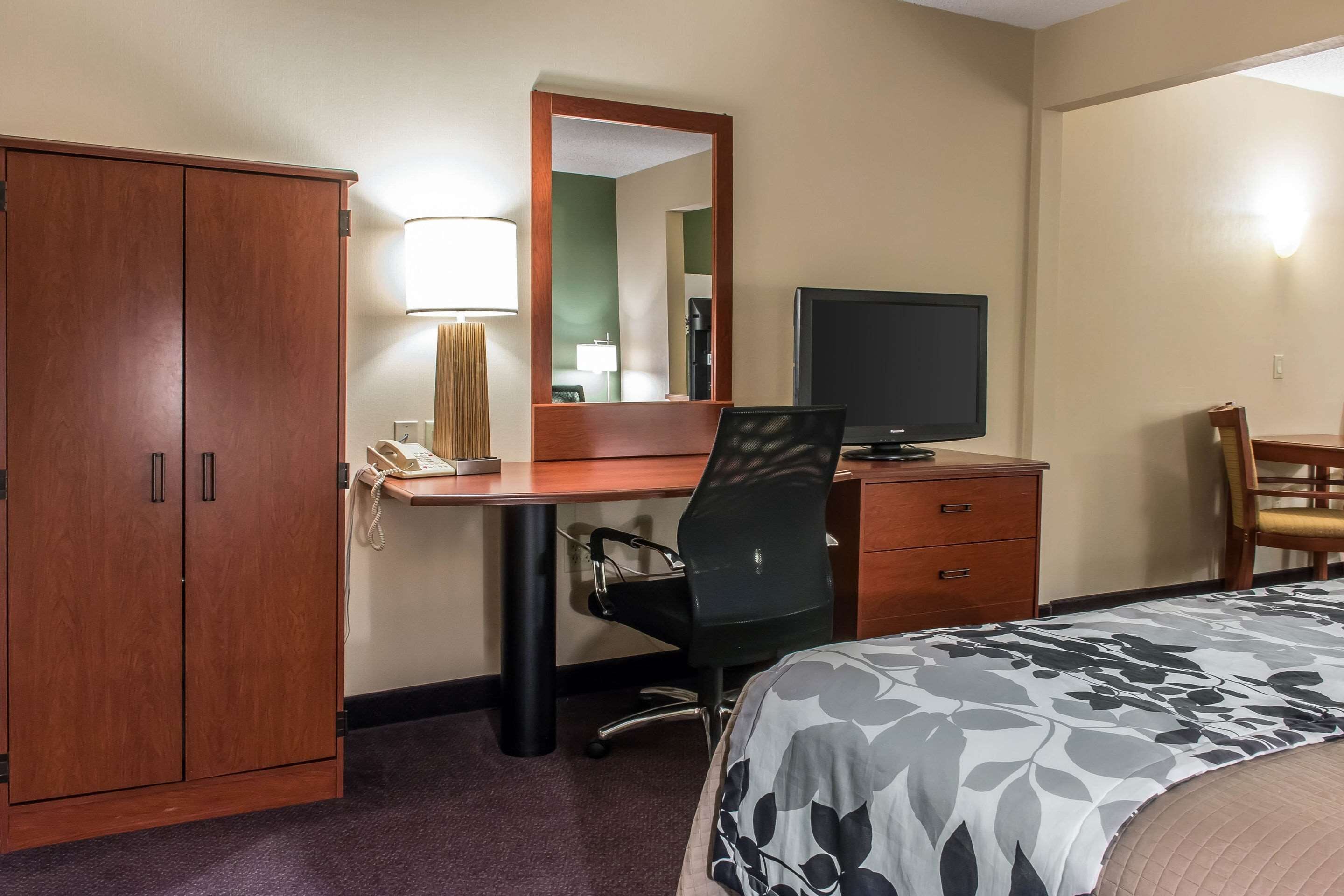 Habitación de hotel con un armario de madera, un escritorio con teléfono, una silla, un televisor y una cama con colcha estampada.