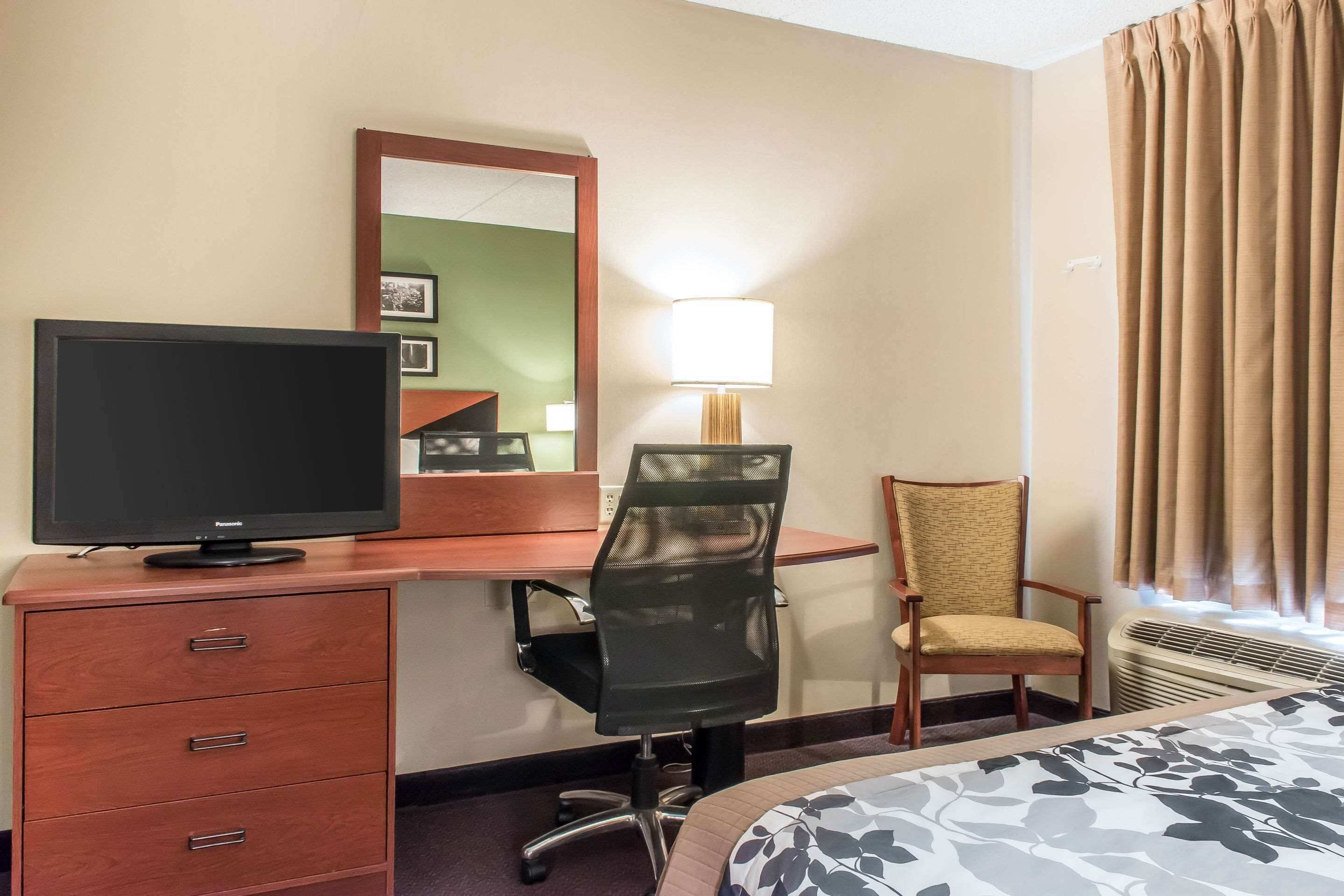 Habitación de hotel con cama, escritorio con monitor de computadora, silla, lámpara y ventana con cortinas.