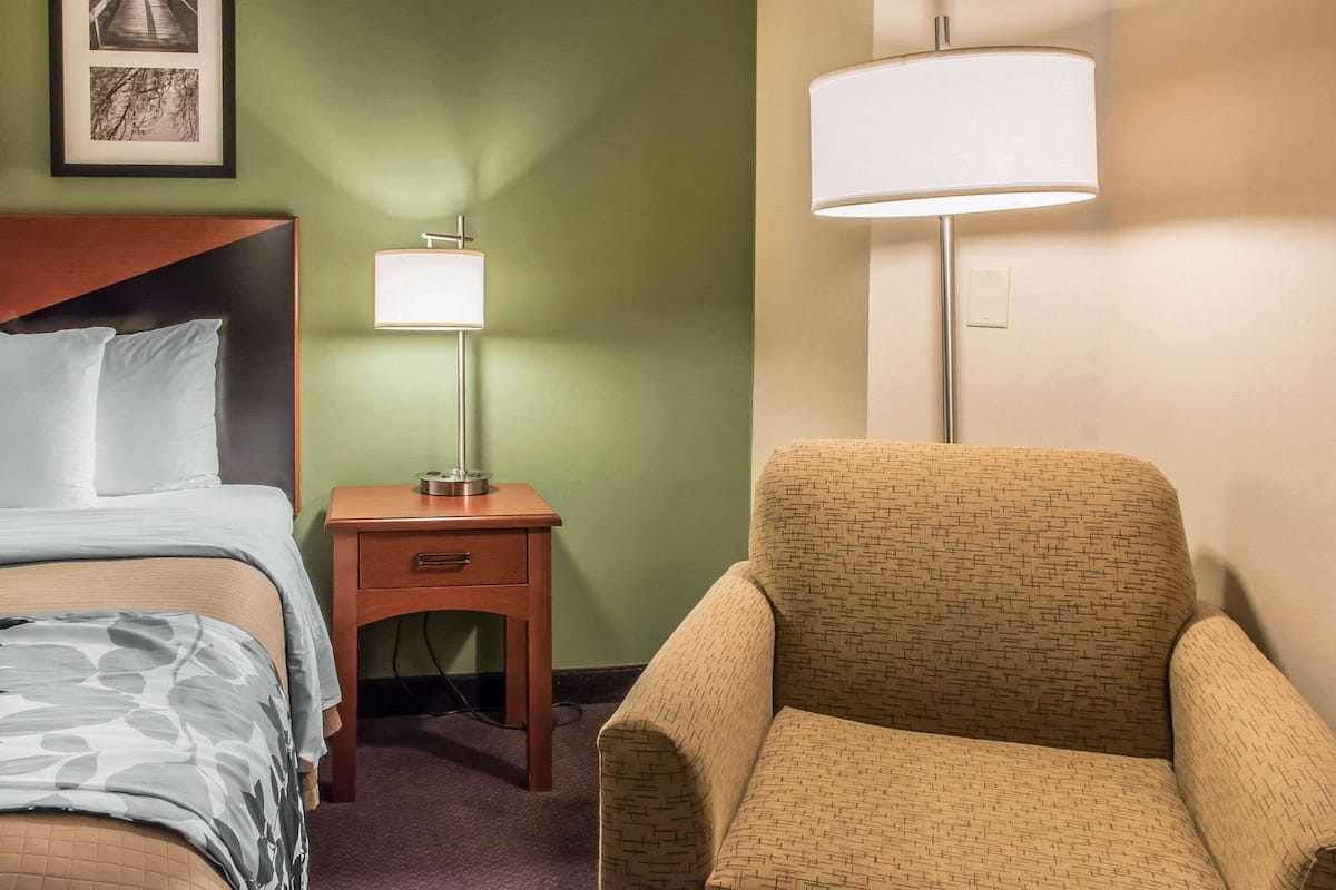 A hotel room with a bed, a nightstand with a lamp, and a patterned armchair.