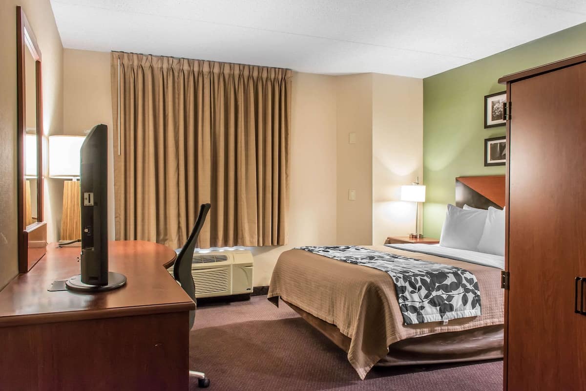 A hotel room with a bed, a desk with a television, an air conditioner, a window with curtains, a nightstand with a lamp, and a door.