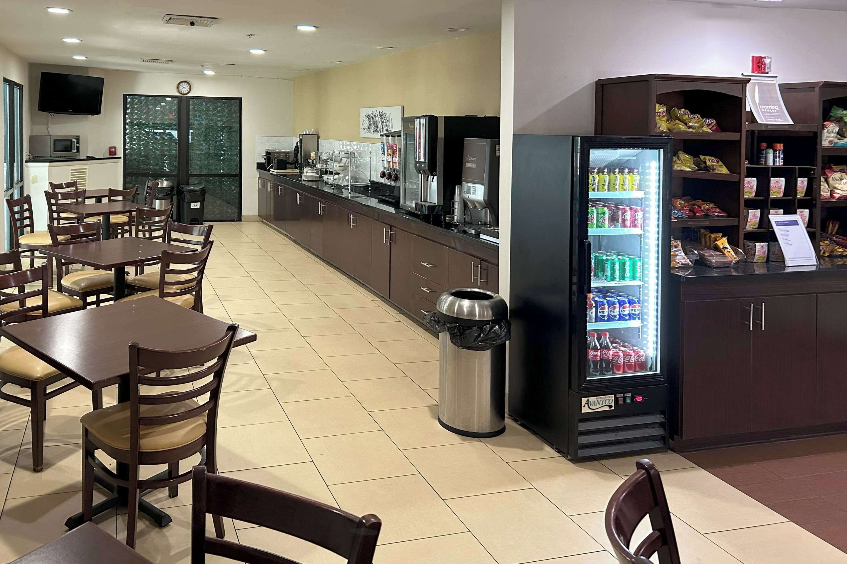 A hotel restaurant with wooden tables and chairs, a coffee machine, a trash can, and a beverage refrigerator.
