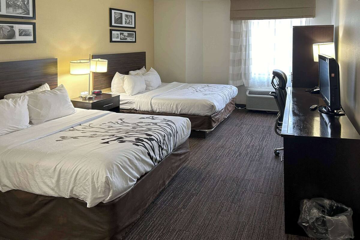 A hotel room with two beds, a desk with a computer, a lamp, and a window with curtains.
