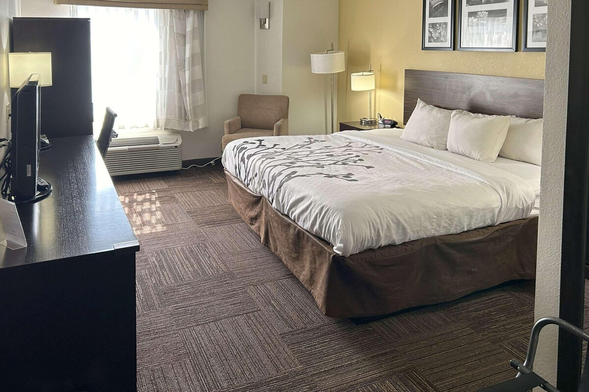 A hotel room with a large bed, a desk with a computer, a chair, a lamp, and a window with curtains.