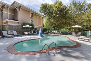 A swimming pool with a brick edge, surrounded by lounge chairs and umbrellas, with a building in the background.