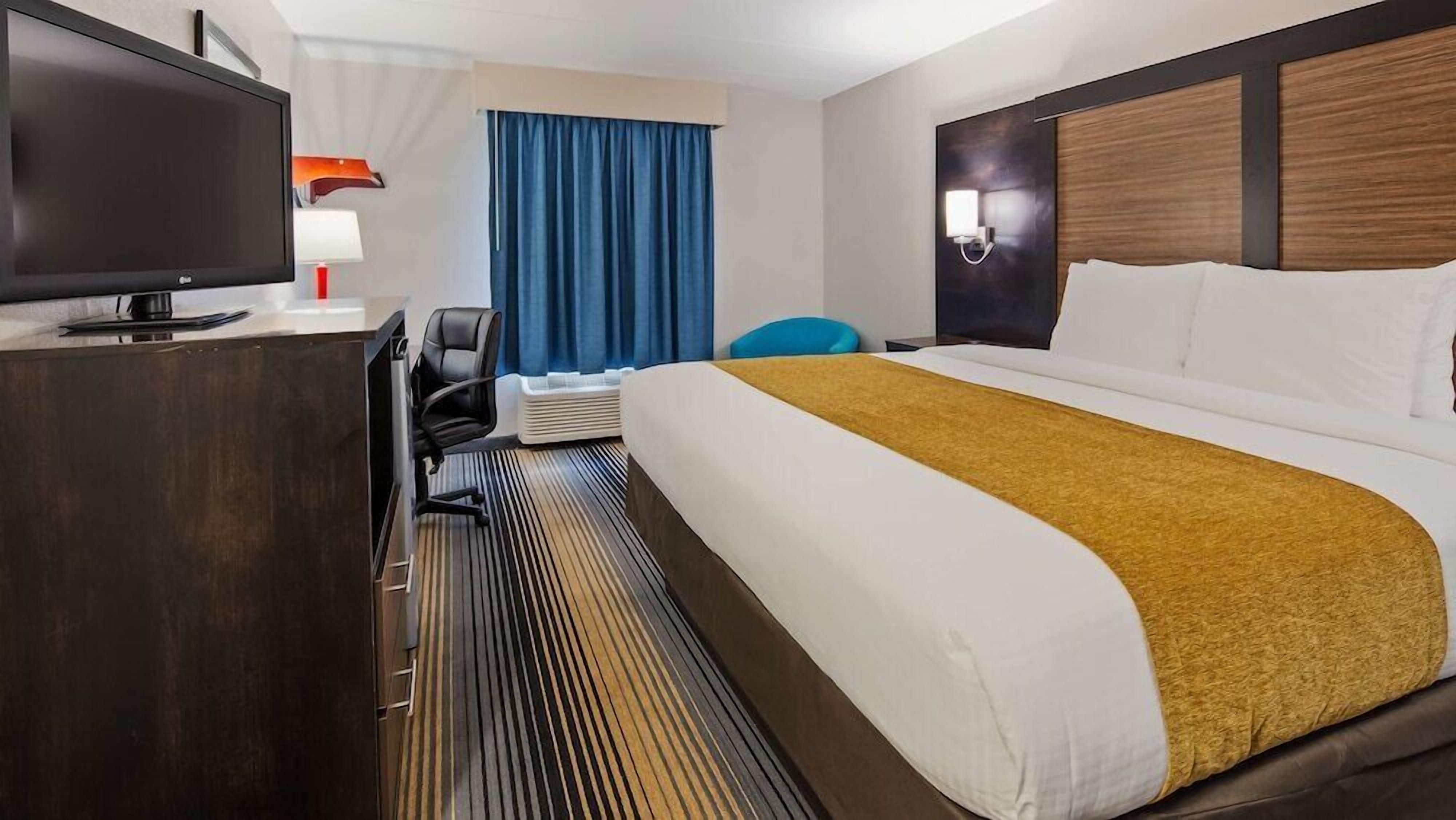 A hotel room with a bed, a desk with a TV, a chair, a window with curtains, and a wall-mounted light.
