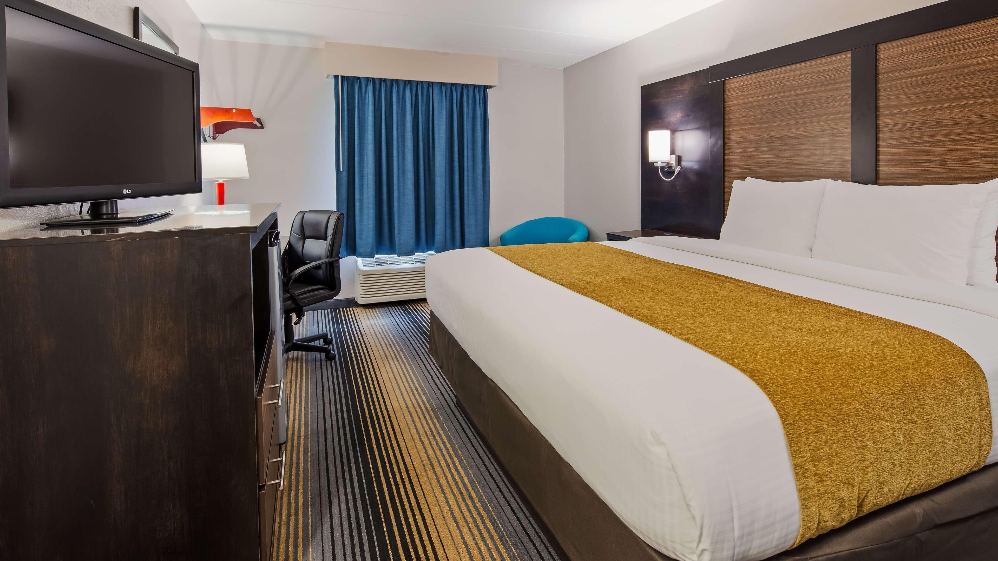 Hotellihuone, jossa on sänky, työpöytä, jossa on televisio, tuoli, ikkuna verhoilla ja seinään kiinnitetty valaisin.