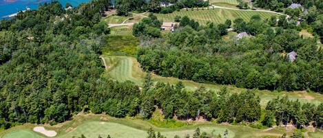 An aerial view of a golf course with multiple holes, surrounded by dense forests and a large body of water.