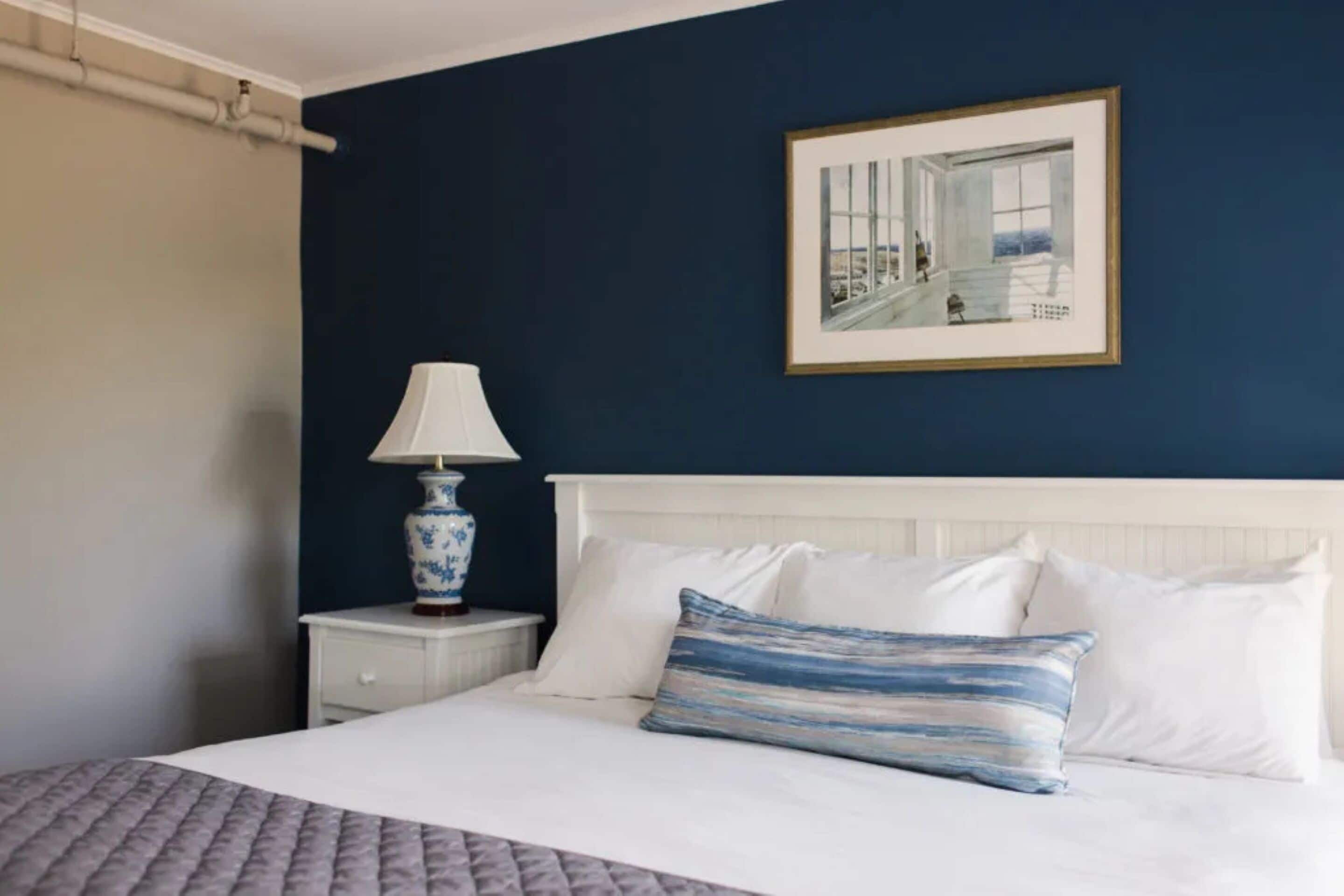 Una cama bien hecha con ropa blanca y una almohada a rayas azules y blancas, una mesita de noche blanca con una lámpara de mesa en un florero azul y blanco, y un cuadro en la pared.