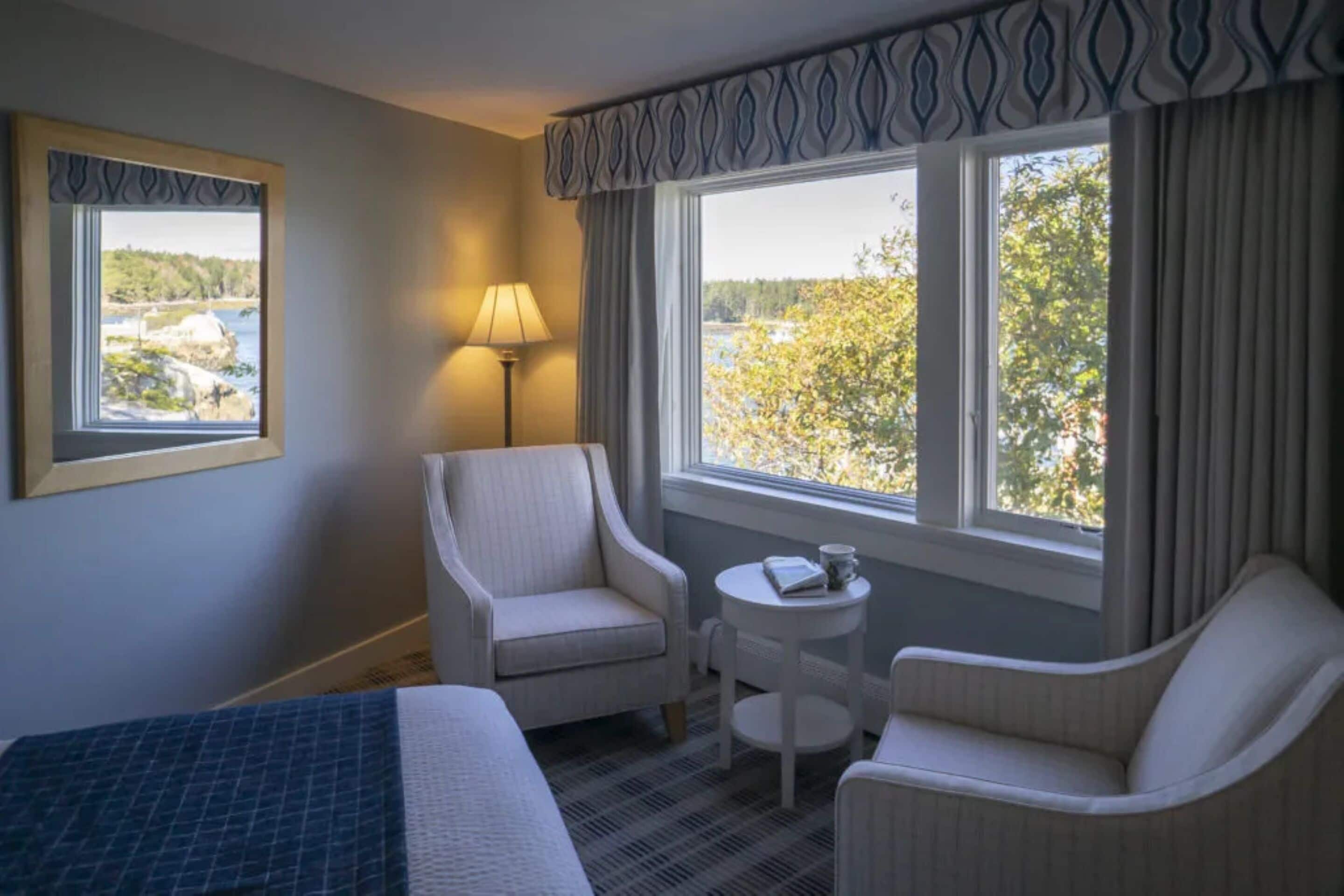 Habitación de hotel con cama, dos sillones, una mesita de noche con lámpara y vistas a un paisaje natural a través de dos ventanas.