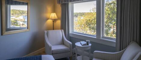 Habitación de hotel con cama, dos sillones, una mesita de noche con lámpara y vistas a un paisaje natural a través de dos ventanas.